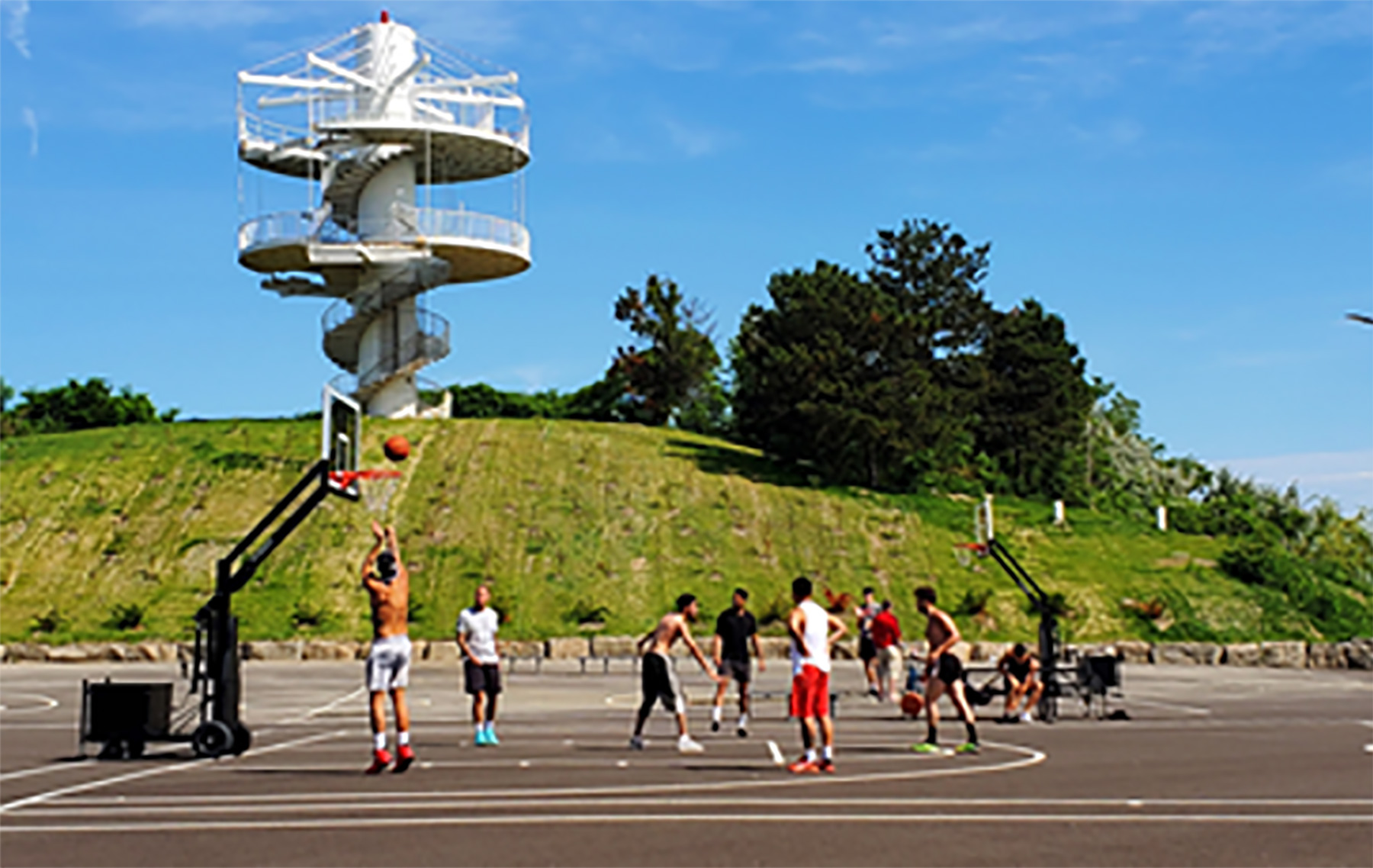 A group of people running and through a basketball on a paved surface with a basketball net. A grassy hill and a tower are in the background. 