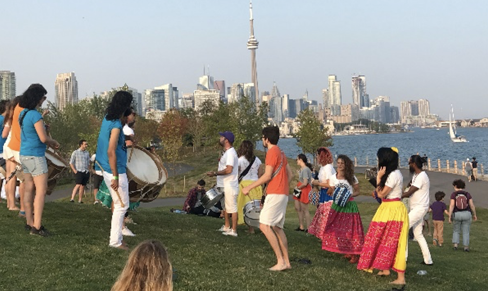 A group of people standing or dancing on a grassy hill with a body of water and a city skyline in the background 