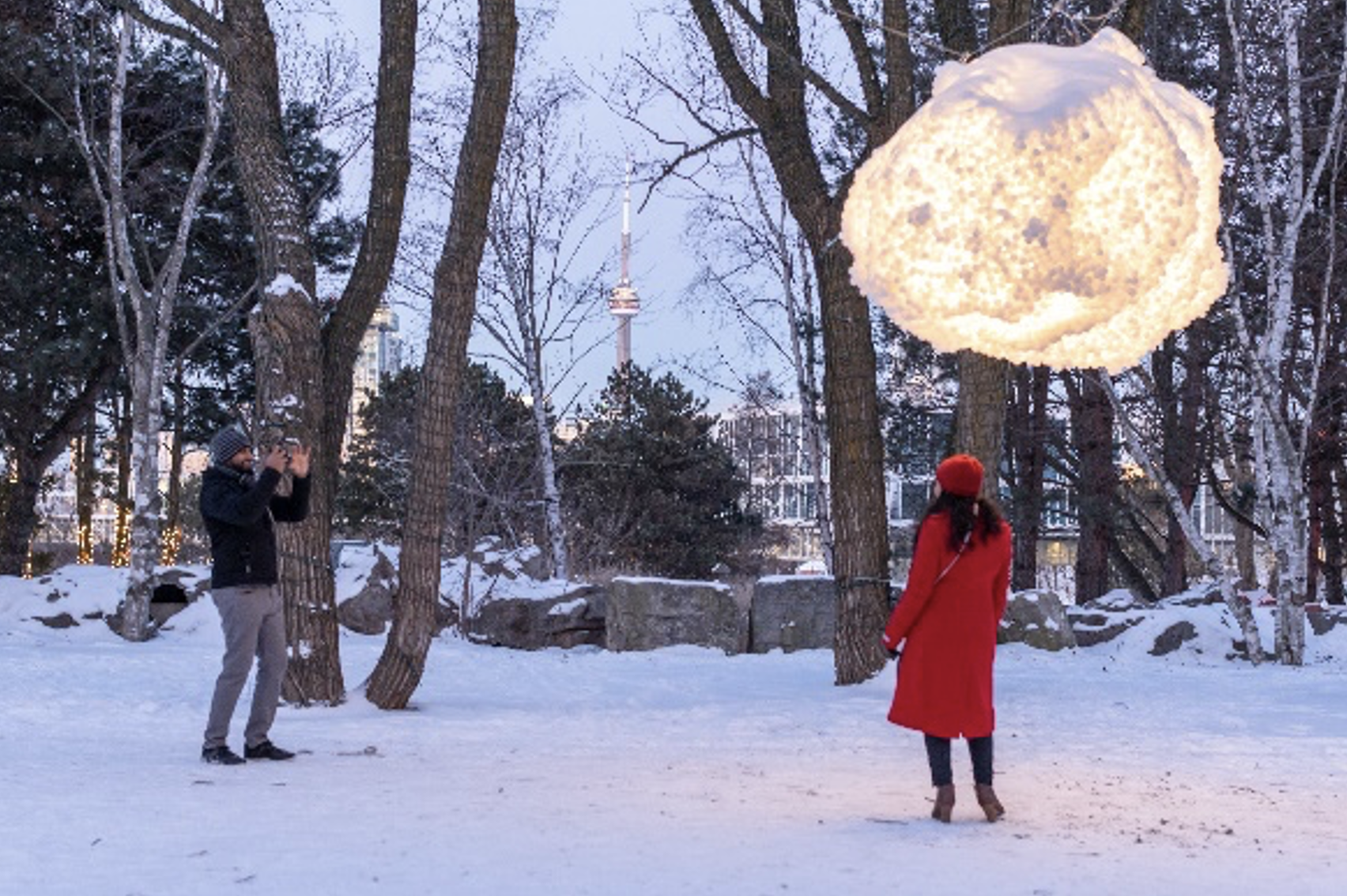 Image of a woman standing beneath a fluffy hanging illuminated light. A man stands away taking her picture. 