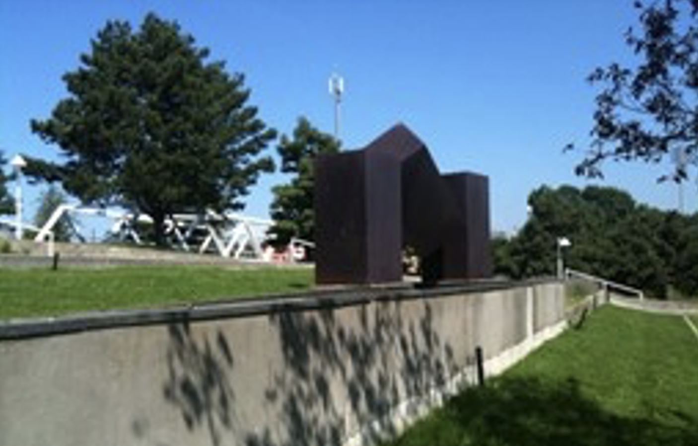Image of an all-black artwork piece on a grassy area above a short concrete wall 

