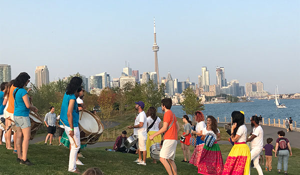 Photo de gens réunis en cercle et jouant du tambour sur un espace vert. Panorama de la ville de Toronto visible en arrière-plan.