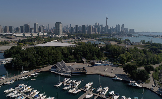 Aerial photo of Ontario Place’s marina, and tree canopy on center island. The CN tower and Lake Ontario is visible in the background.