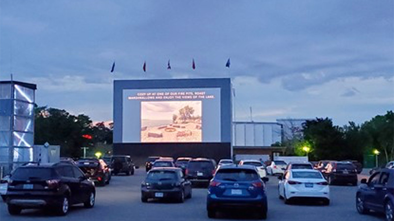 Cars parked facing a large outdoor movie screen at dusk.