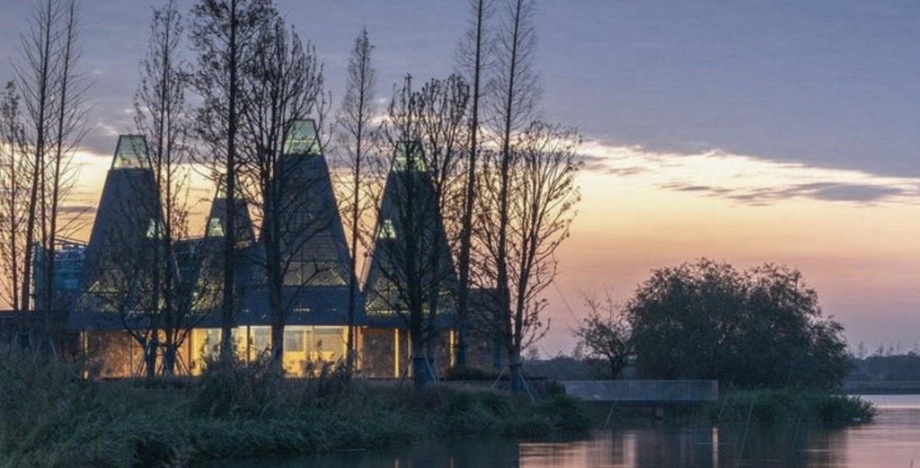 Picture of a building with skinny triangular roofs amongst trees along a body of water with the sun setting