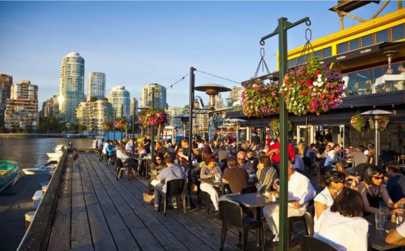 People sitting at an outdoor restaurant along a body of water to the left and a building to the right. Tall city buildings in the background.