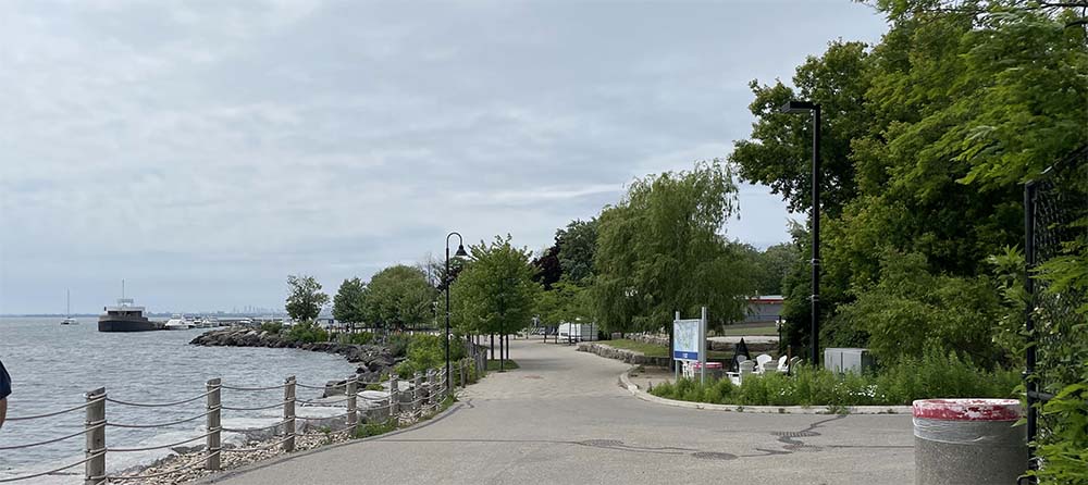 Image of a body of water (Lake Ontario) on the left with the shoreline and a paved pathway in the center of the image and trees on the right.